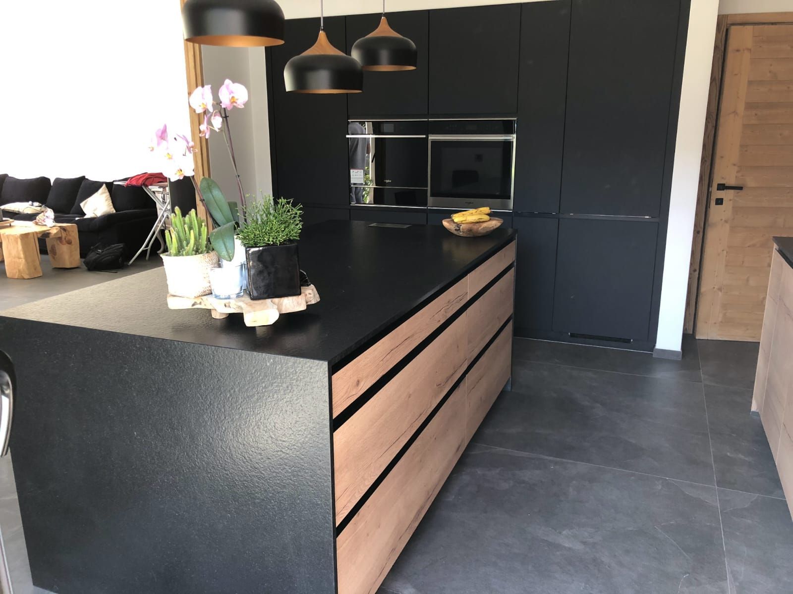 Un îlot de cuisine moderne avec des surfaces noires mates et des tiroirs en bois clair, agrémenté de suspensions lumineuses et de plantes en pot.