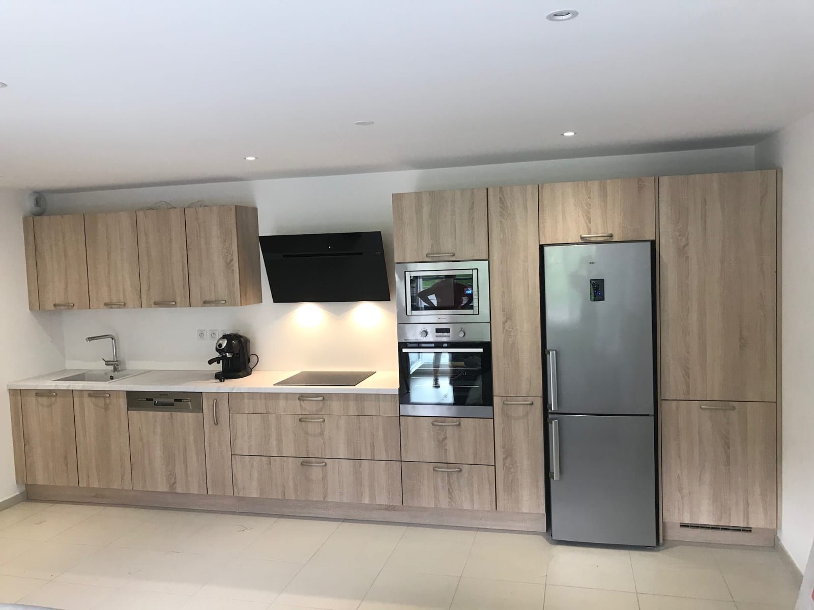 Une cuisine moderne avec des armoires en bois clair, un réfrigérateur en acier inoxydable, un four et une hotte aspirante noire.