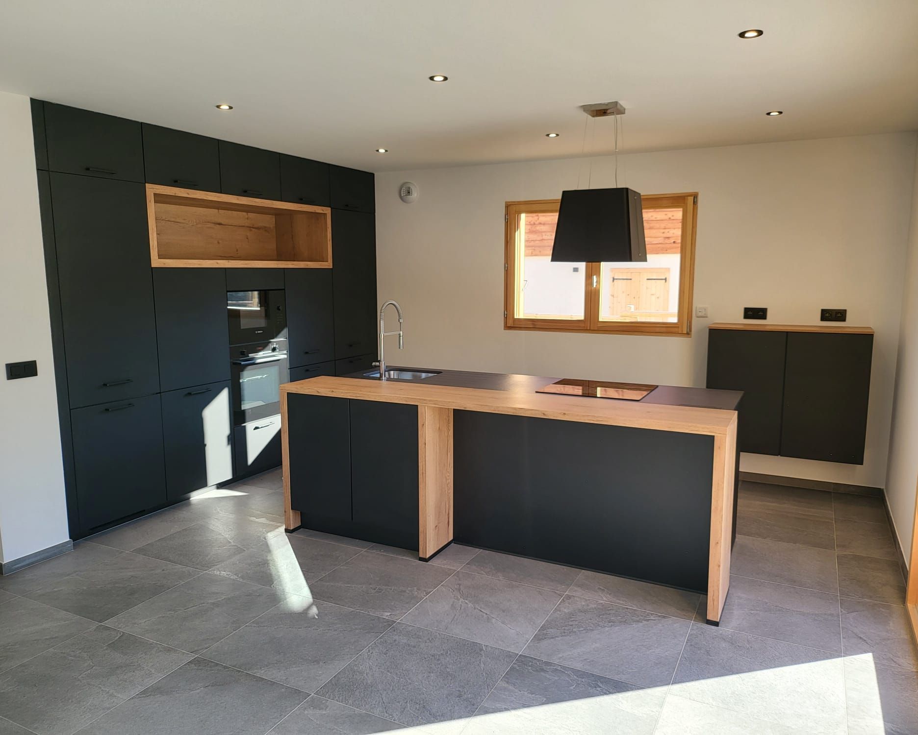 Une cuisine moderne avec des armoires noir mat, un îlot central avec un plan de travail en bois, un sol en carrelage de pierre et une fenêtre à encadrement en bois.