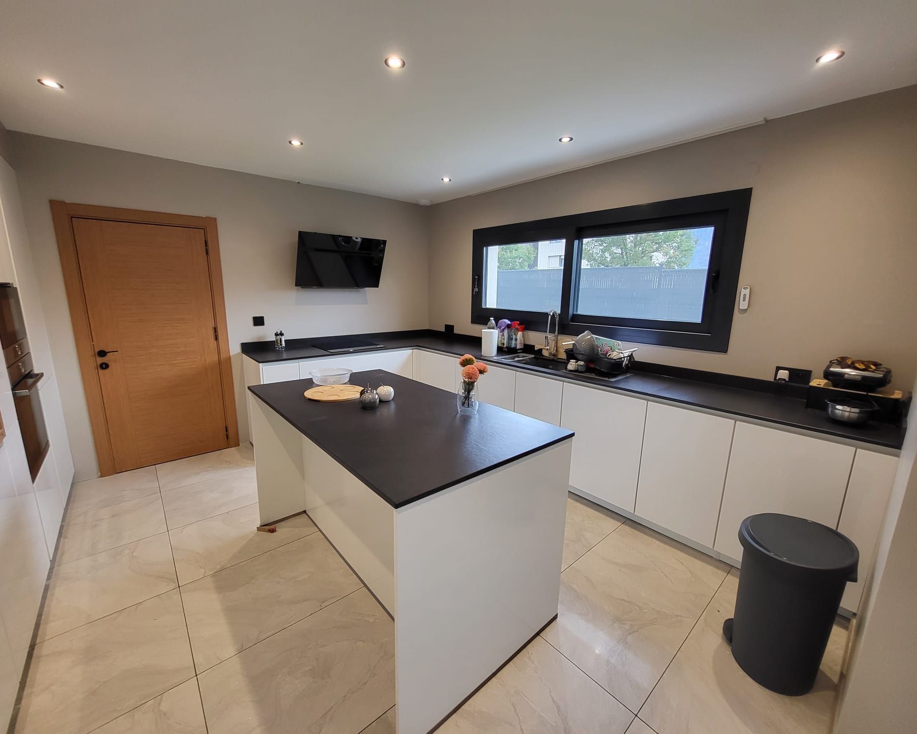 Une cuisine moderne et lumineuse avec des armoires blanches, un îlot et des comptoirs noirs, une porte en bois et un sol carrelé.