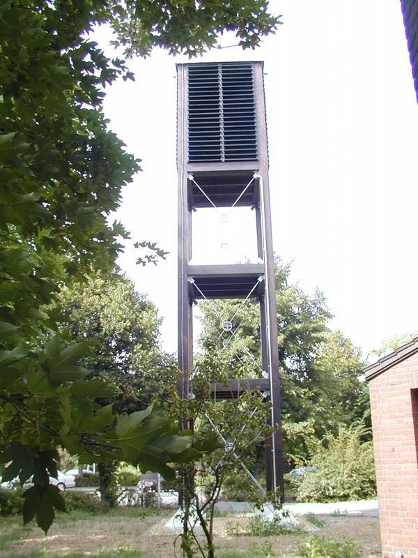 Ein Uhrturm ist von Bäumen und einem Backsteingebäude umgeben