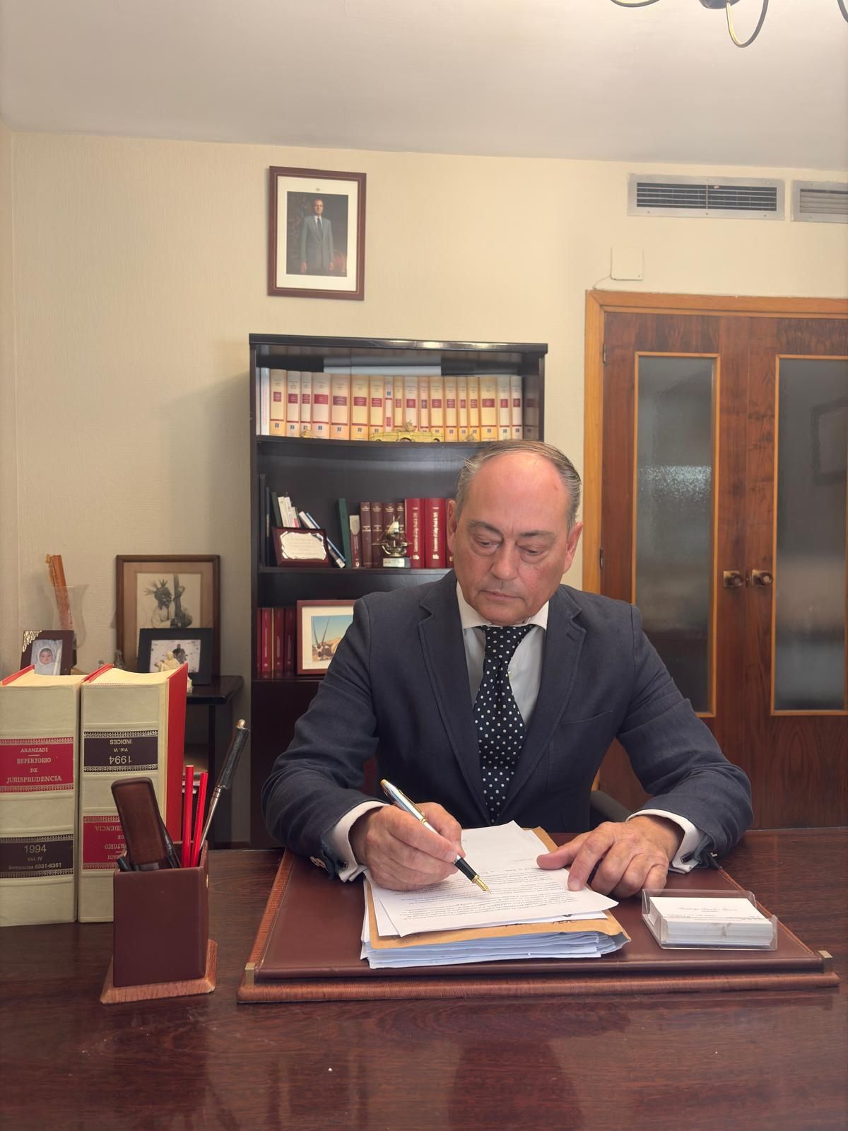 Un hombre de traje escribe en un escritorio de oficina. Se ven una estantería y fotos enmarcadas.