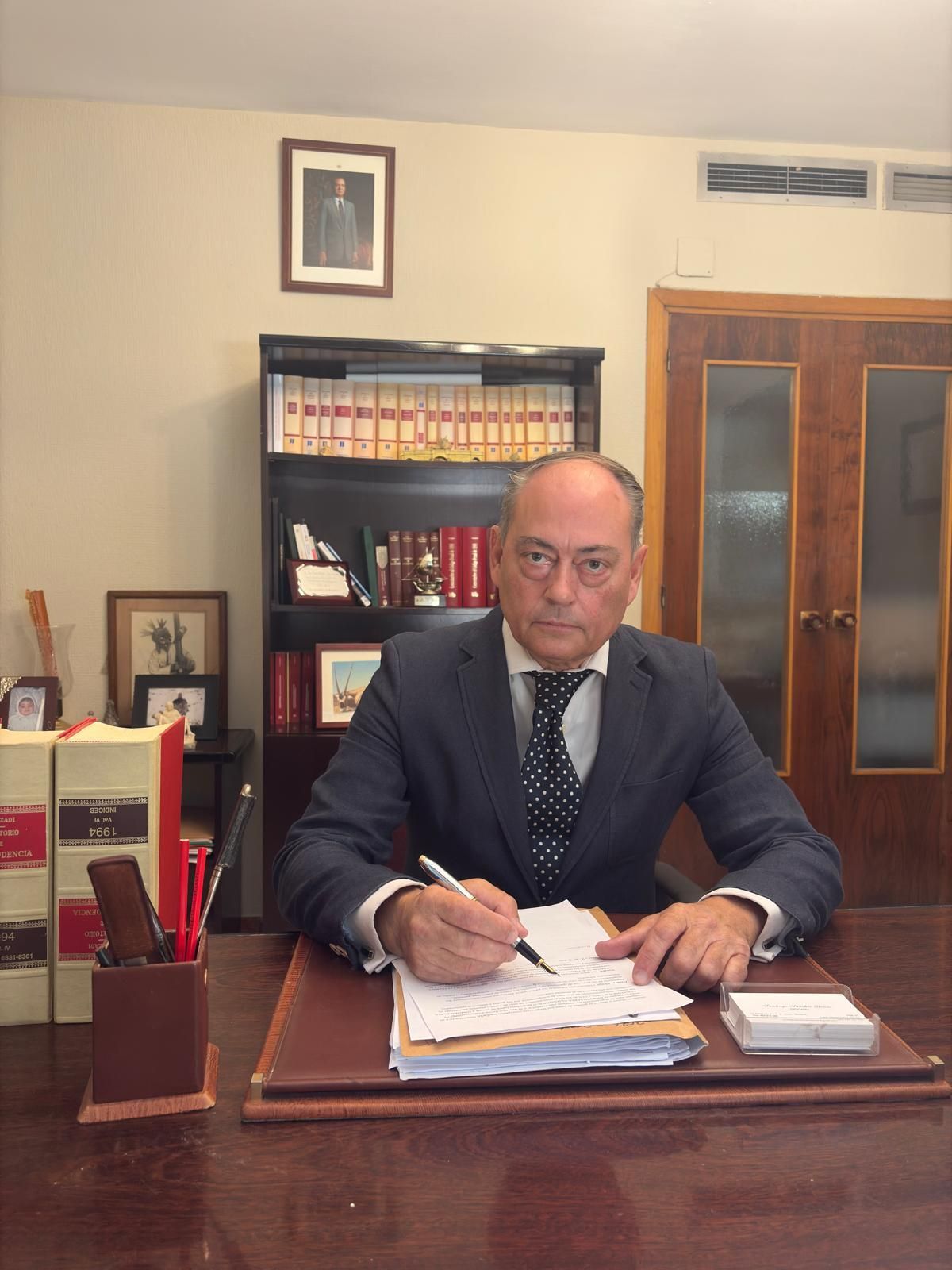 Un hombre de traje está sentado en un escritorio, escribiendo. Está en una oficina con estanterías y fotos.