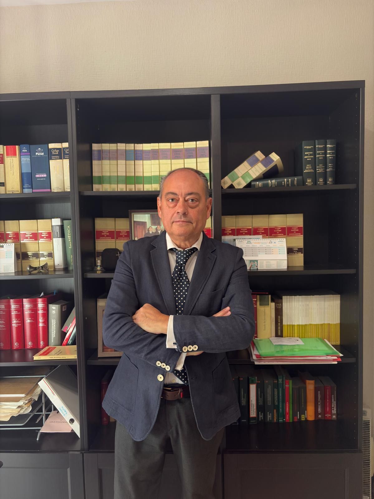 Hombre con blazer y brazos cruzados delante de una estantería llena de libros.