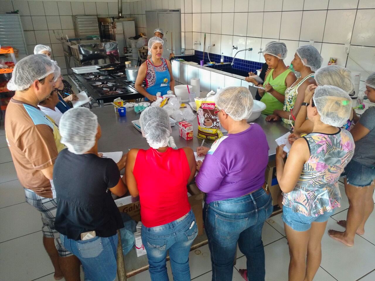 Eine Gruppe von Menschen mit Haarnetzen versammelte sich um einen Arbeitstisch in einem Raum zur Lebensmittelzubereitung.
