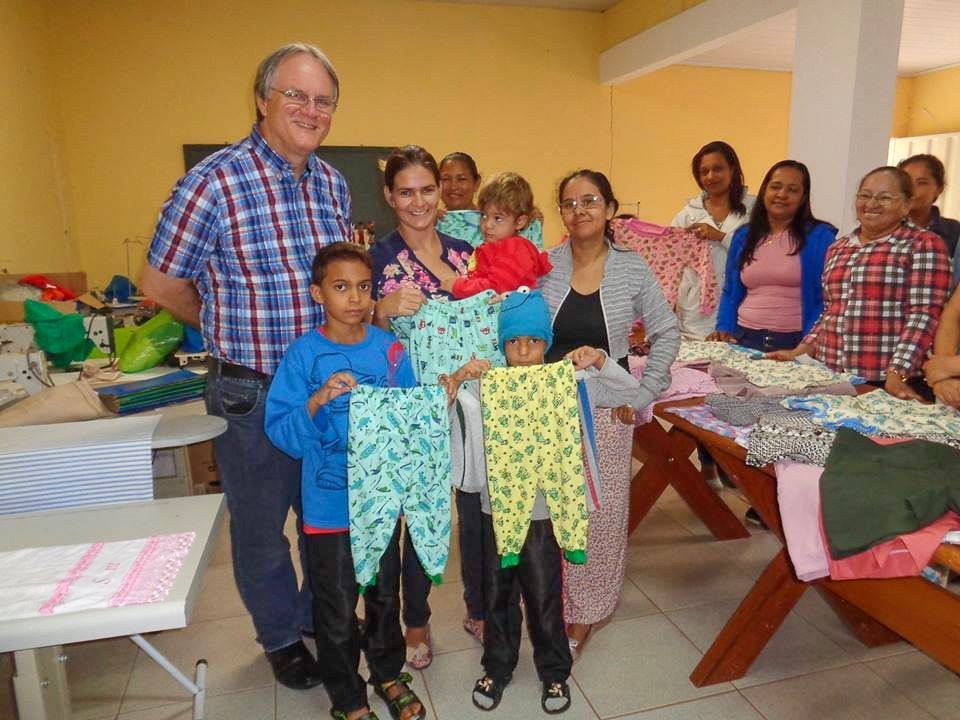 Eine Gruppe hält bunte Babykleidung und Decken in einem Raum mit Tischen und gefalteter Wäsche.