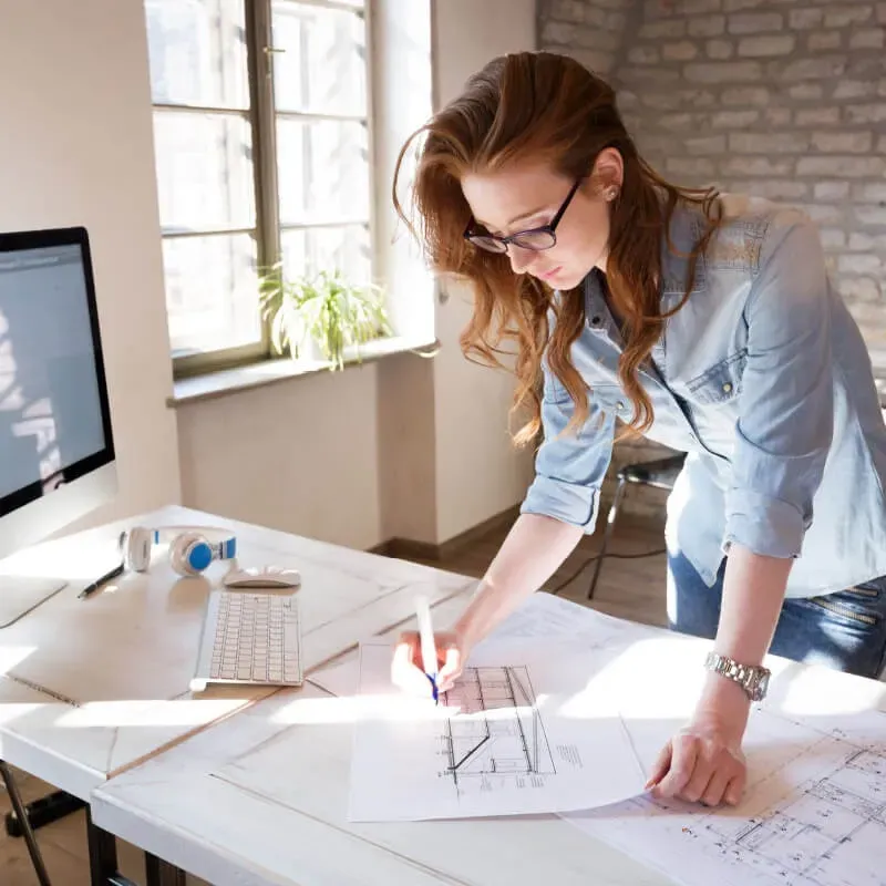 Mujer con cabello rojo y gafas trabajando en planos en un escritorio con una computadora y auriculares.