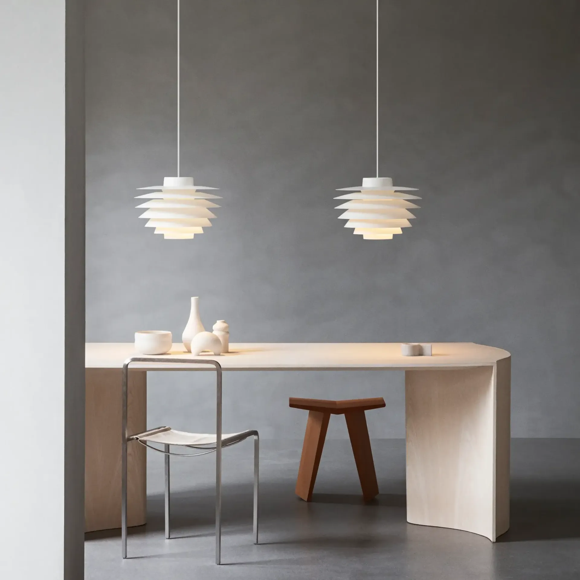 Dos lámparas colgantes blancas iluminan una mesa de comedor con silla y taburete. Pared gris.