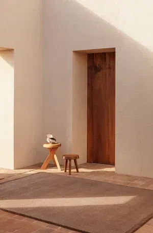 Habitación minimalista con puerta de madera, dos taburetes y una pequeña escultura en uno de ellos. Paredes en cálido beige.