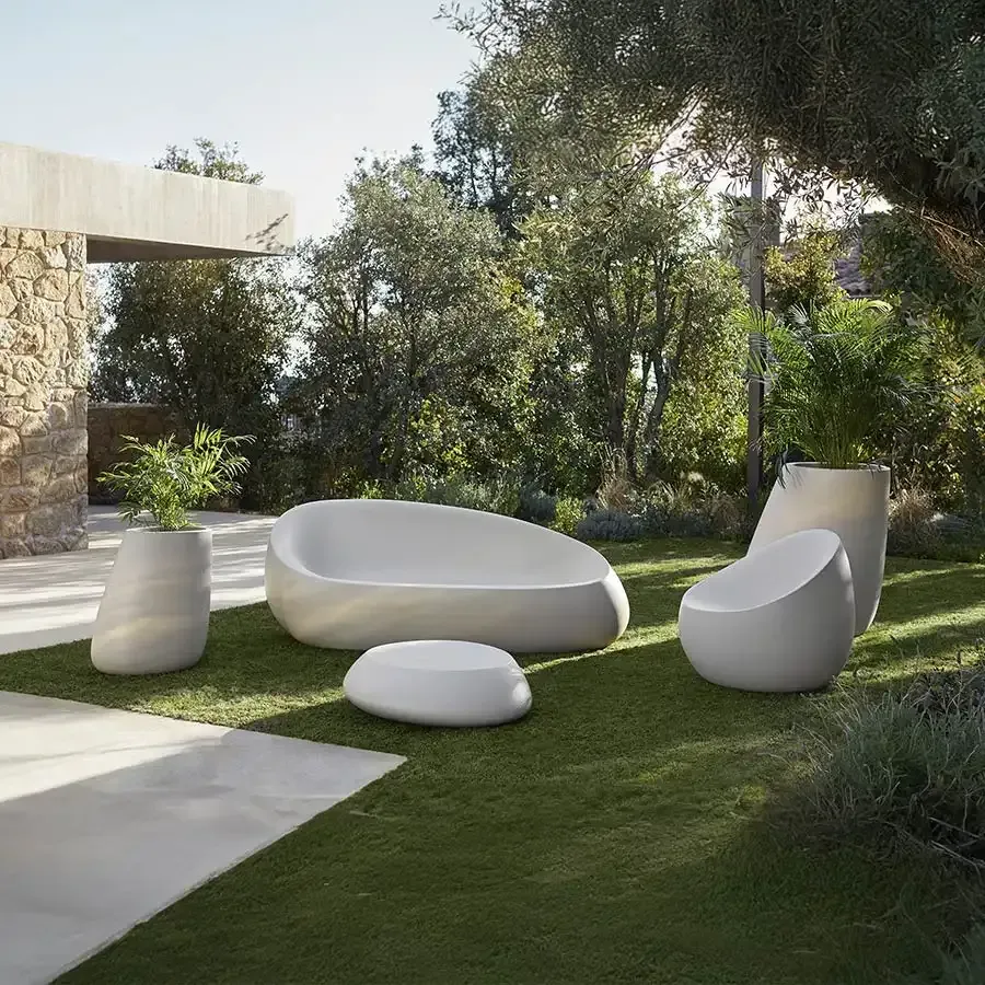 Muebles de exterior situados sobre un césped: sofá blanco, silla y jardineras cerca de un muro de piedra.
