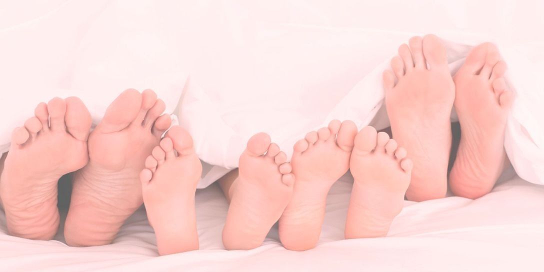Varios pares de pies descalzos asomando por debajo de mantas blancas en una cama rosa.