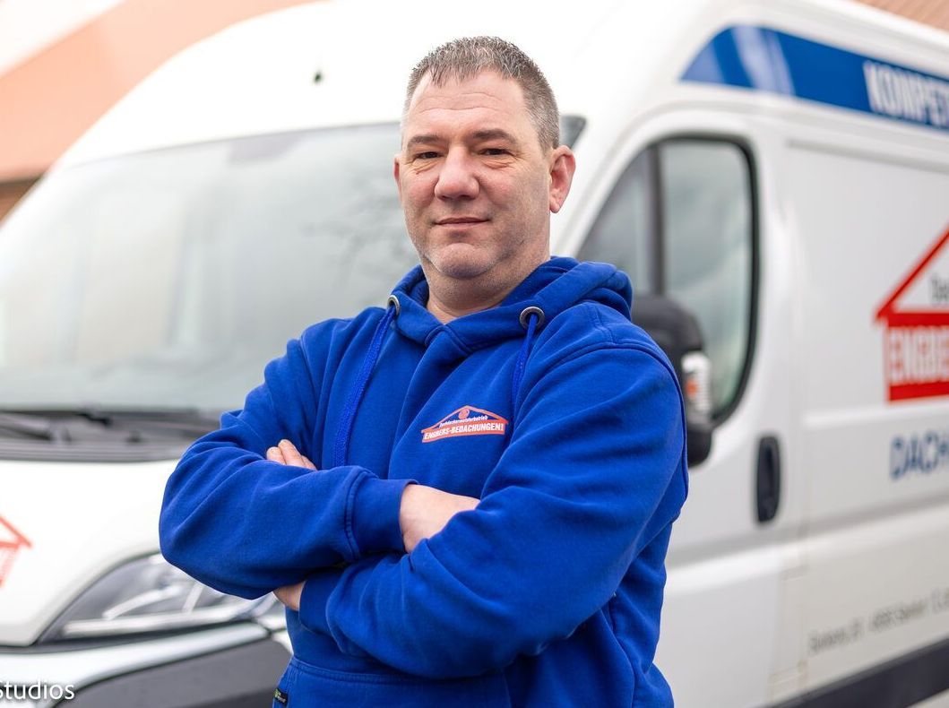 Ein Mann in einem blauen Sweatshirt steht mit verschränkten Armen vor einem weißen Lieferwagen.