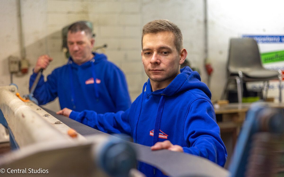 Zwei Männer in blauen Sweatshirts arbeiten in einer Fabrik an einer Maschine.