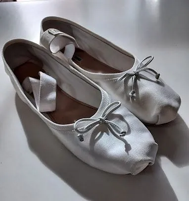 Un par de bailarinas blancas con lazos están sobre una mesa blanca.