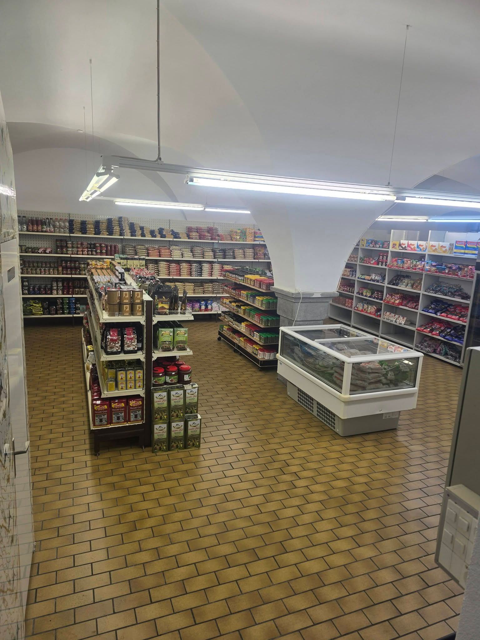 Innenansicht eines ruhigen Lebensmittelgeschäfts mit Regalen voller Produkte, einer Gefriertruhe und einem markanten Fliesenboden.
