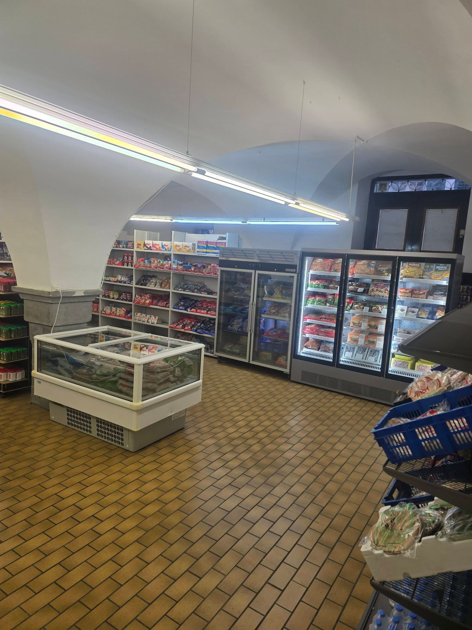 Innenansicht eines kleinen Lebensmittelgeschäfts mit zentralem Gefrierschrank, Kühlschränken mit Glastüren und Regalen mit Lebensmitteln unter gewölbten Wänden.
