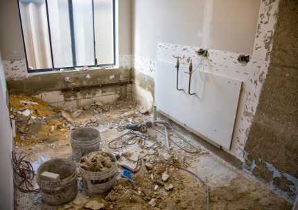 Salle de bain en rénovation : débris sur le sol, tuyaux exposés, murs non finis, seaux, fenêtre et outils.