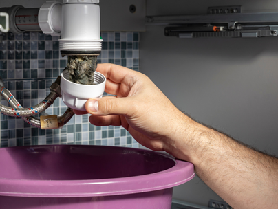 Une main dévisse un siphon d'évier rempli de saleté au-dessus d'un seau violet dans une salle de bain.