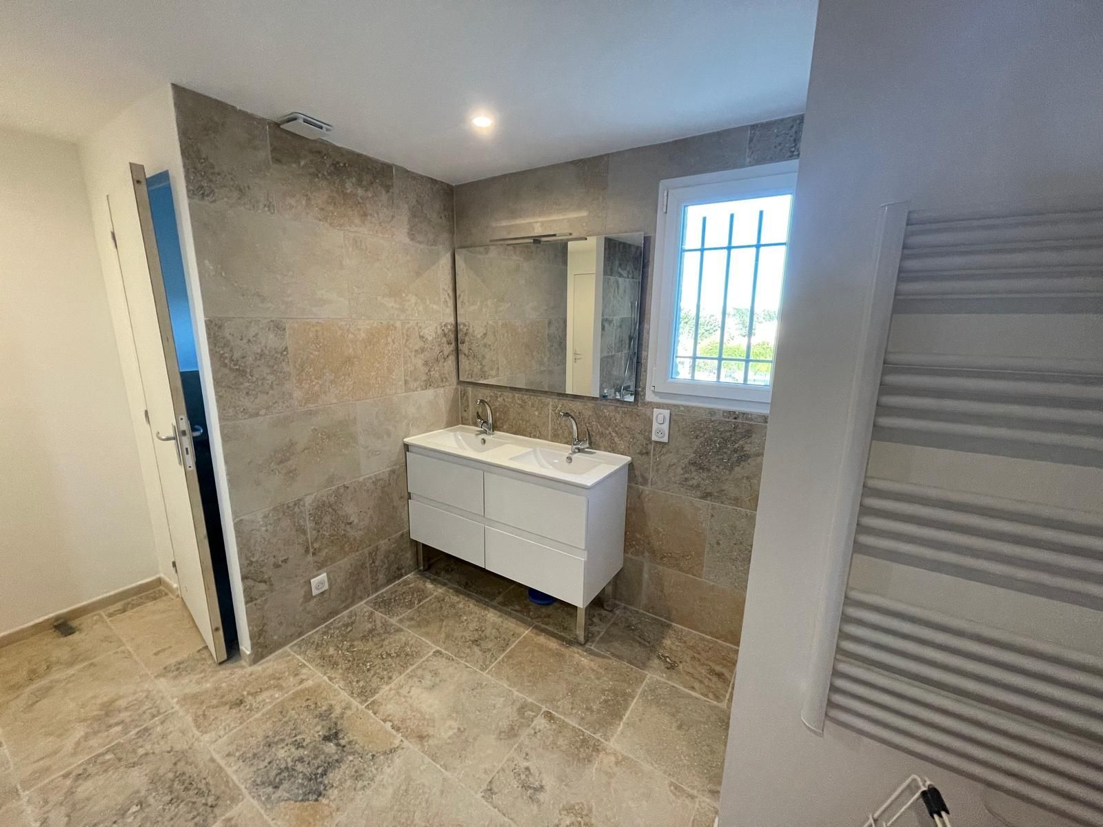 Salle de bain avec double vasque, murs/sol carrelés, fenêtre et porte.