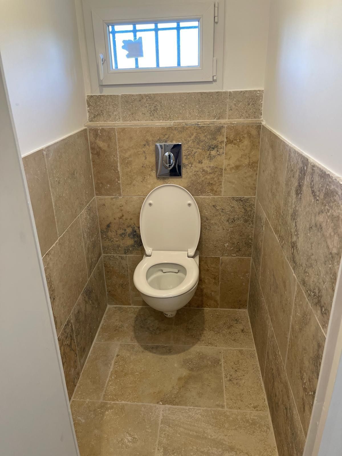 Plomberie sous évier avec siphon et tuyaux chromés contre un mur endommagé et un fond carrelé.