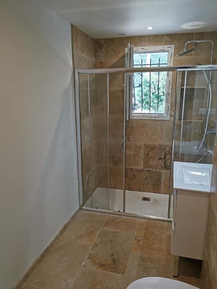 Plombier utilisant une clé pour travailler sur un tuyau chromé sous un évier dans une pièce carrelée blanche.