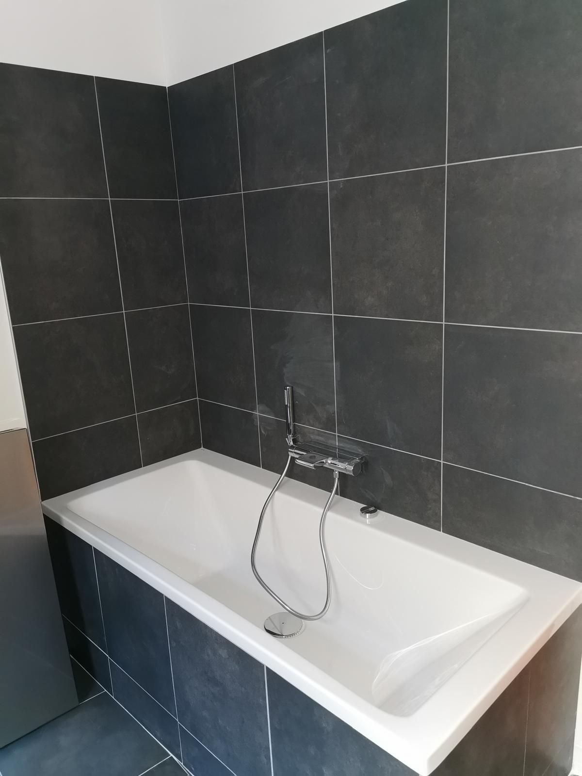 Baignoire blanche encastrée dans un entourage en carrelage foncé, avec accessoire de douche.