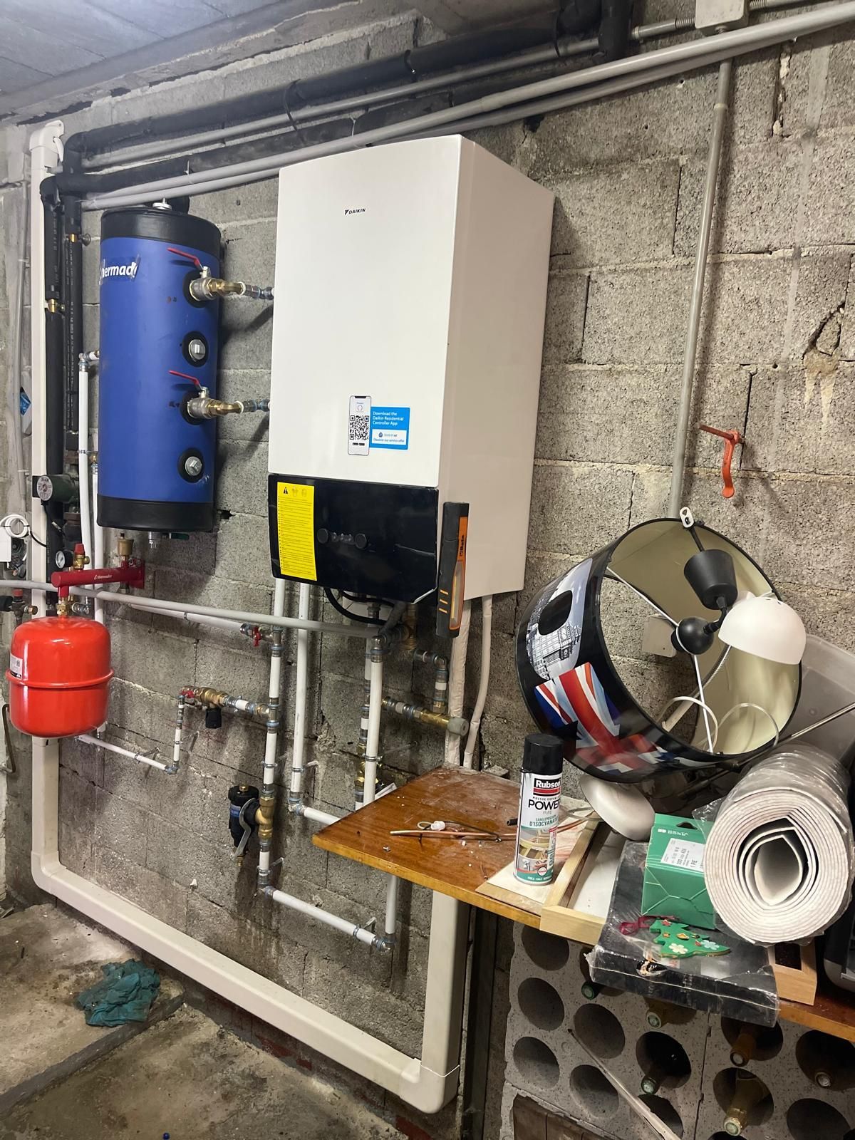 Plomberie sous évier avec siphon et tuyaux chromés contre un mur endommagé et un fond carrelé.