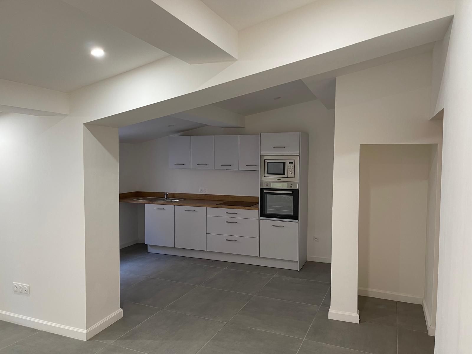Cuisine vide et lumineuse avec armoires, plan de travail et électroménagers blancs. Carrelage gris au sol.