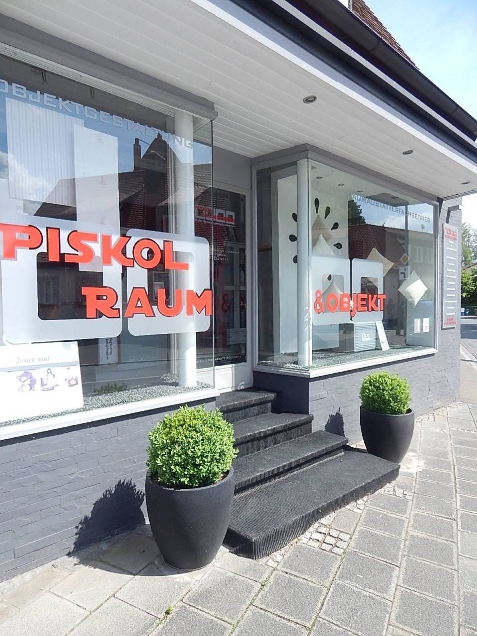 Außenansicht eines Ladens mit Glasfenstern. „PISKOL RAUM“ in Rot. Grüne Topfpflanzen flankieren die Eingangstreppe.