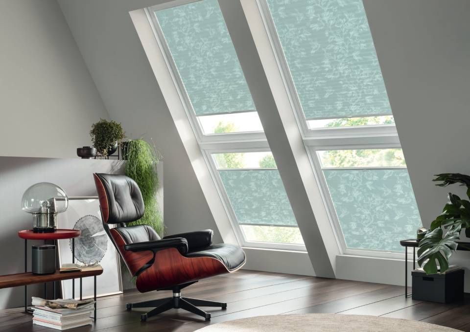Modernes Dachzimmer mit schrägen Fenstern, blaugrün gemusterten Jalousien, einem schwarz-braunen Sessel und Pflanzen.