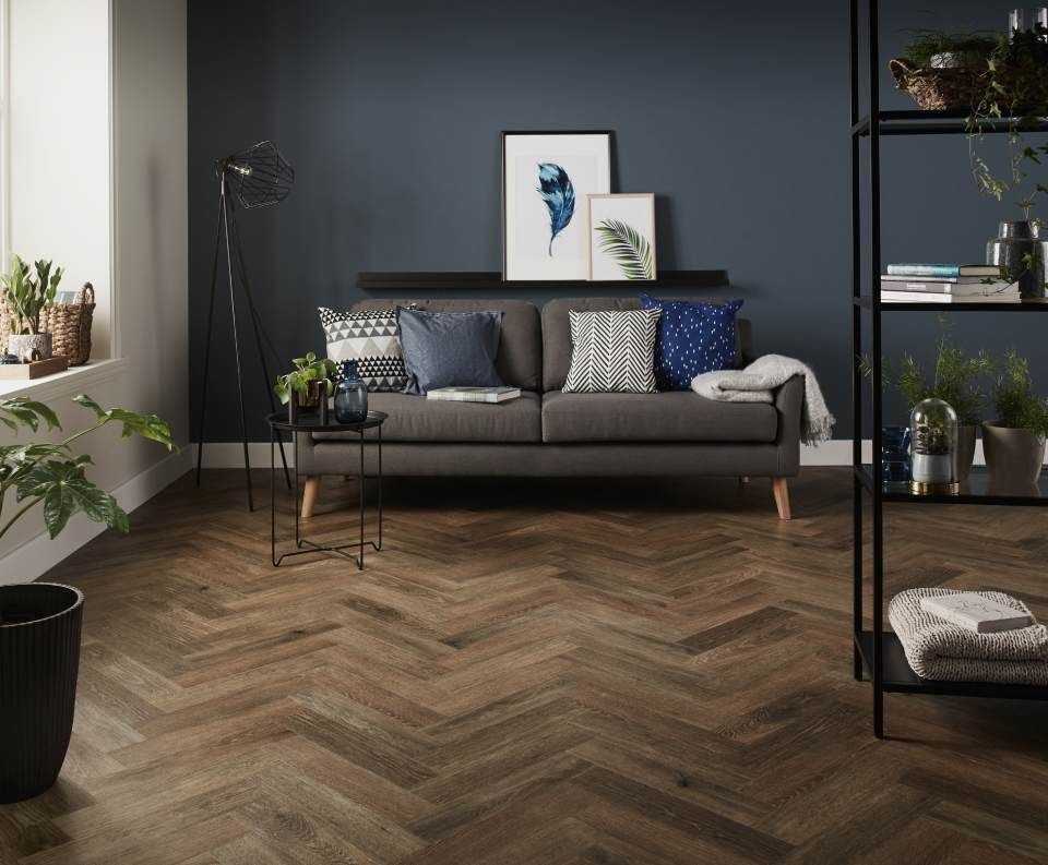 Wohnzimmer mit Fischgrätholzboden, grauem Sofa vor einer dunkelblauen Wand und schwarzen Regalen.
