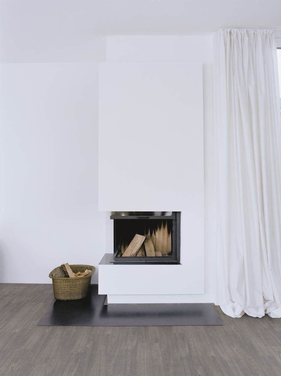Moderner weißer Kamin mit Brennholz, Korb auf Holzboden. Weiße Vorhänge rechts.