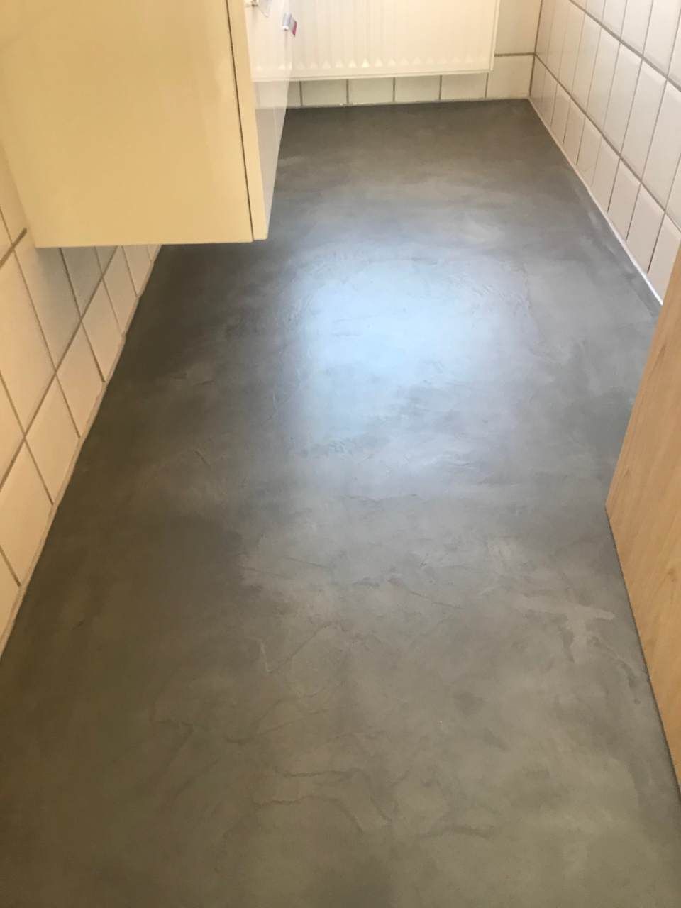 Grauer Betonboden in einem Badezimmer mit weiß gefliesten Wänden und einer Holztür.
