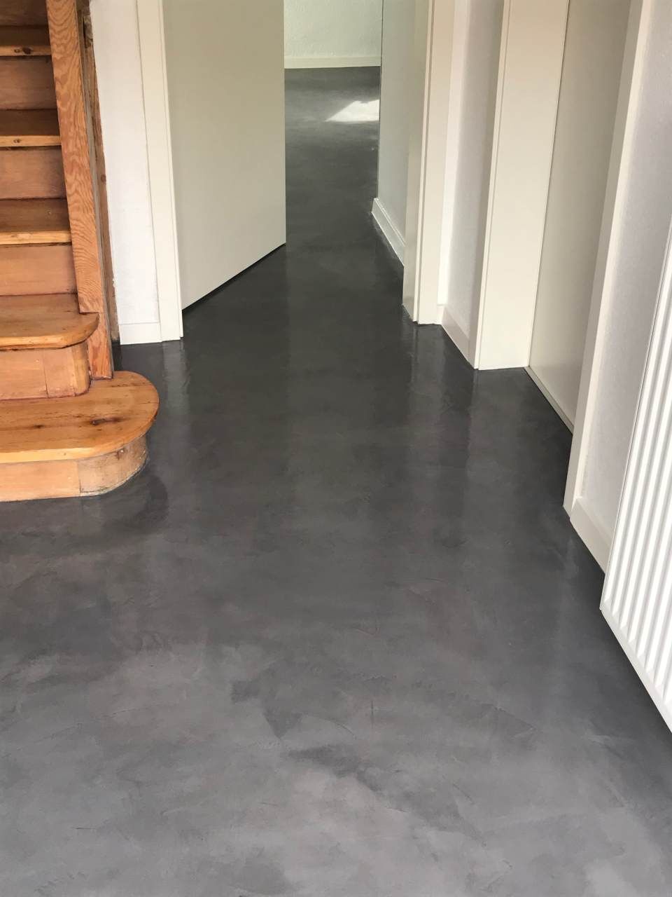 Grauer, polierter Betonboden in einem Flur mit weißen Wänden und einer Holztreppe.