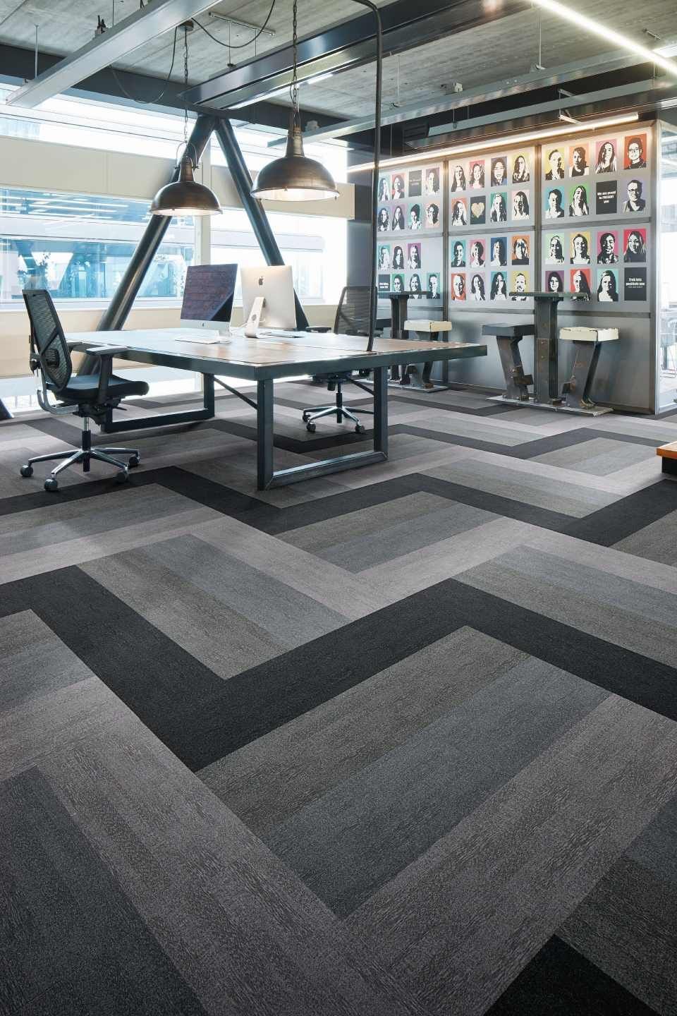 Büroraum mit geometrischem Teppichmuster, Schreibtischen, Stühlen und einer Wand mit gerahmten Fotos.
