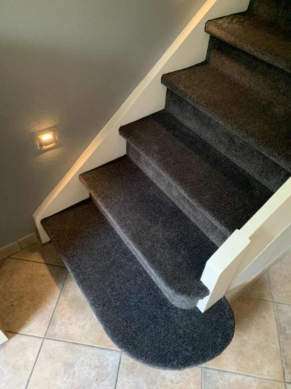 Mit dunklem Teppich ausgelegte Treppe mit weißen Zierleisten vor einer grauen Wand, auf Fliesenboden.