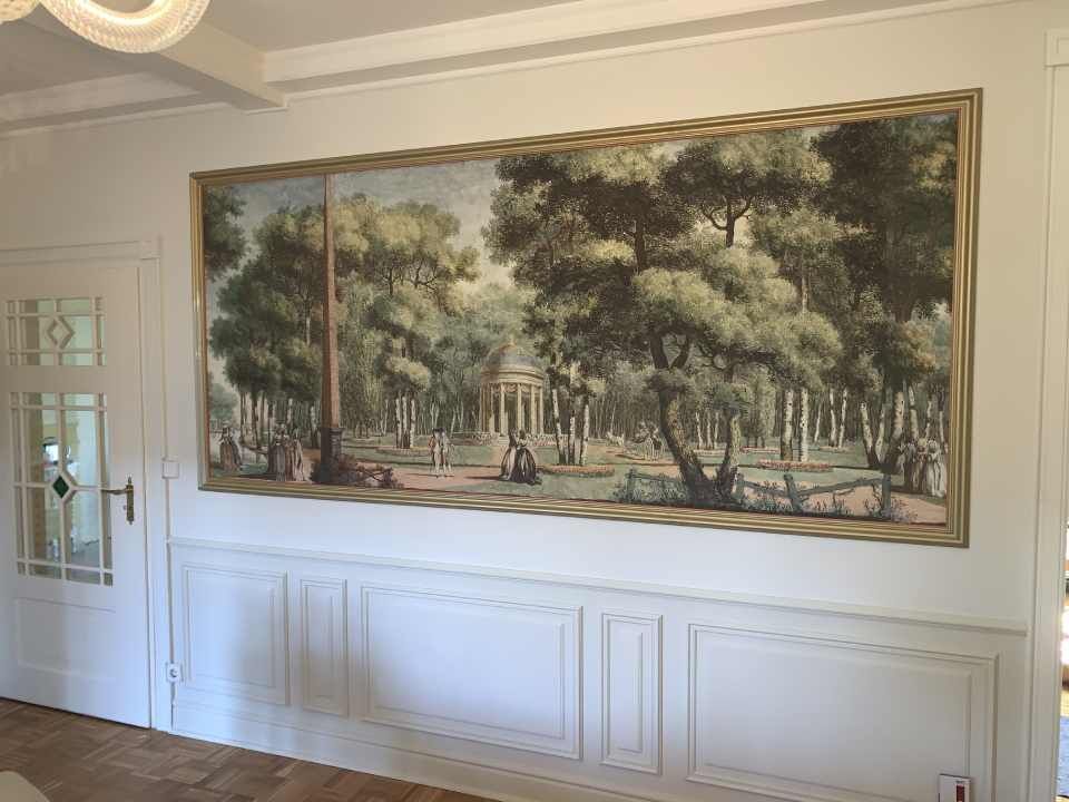 Großes gerahmtes Landschaftswandbild mit Bäumen und Figuren in einem Flur mit weiß getäfelten Wänden und einer Tür.