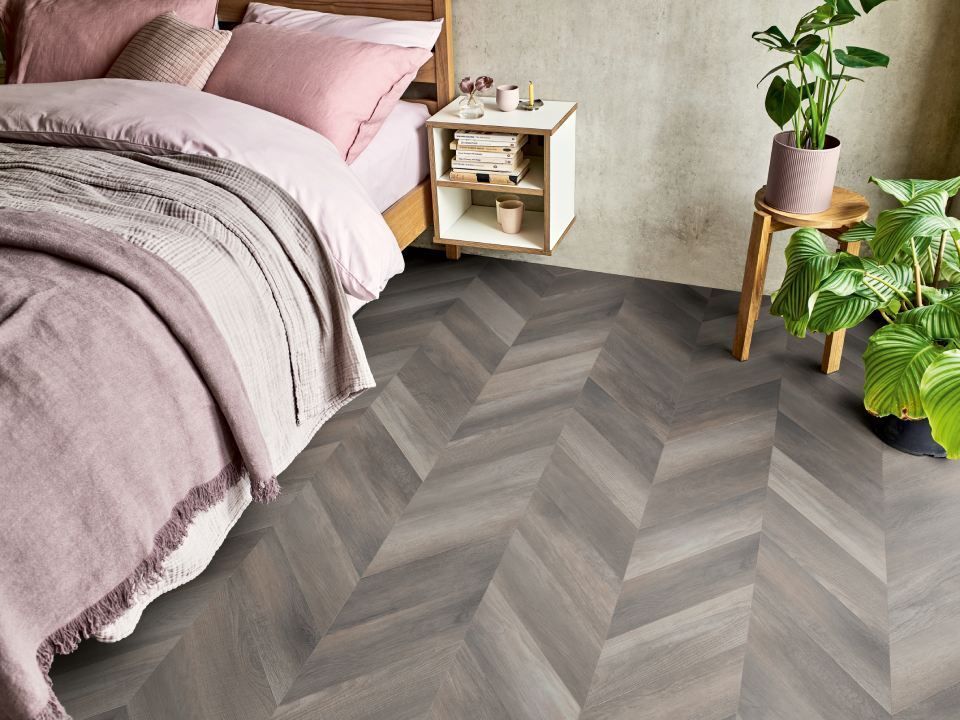 Schlafzimmer mit Boden in Fischgrät-Holzoptik, Bett mit rosa Bettwäsche und Decke.