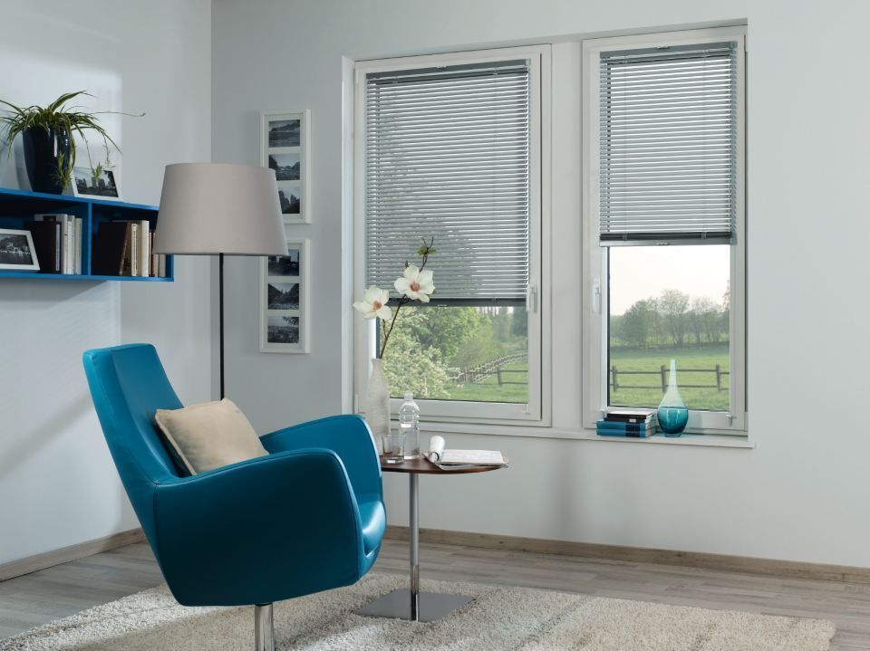Blauer Sessel, Lampe und Regale in einem Wohnzimmer mit Jalousien und Blick auf eine grüne Wiese.