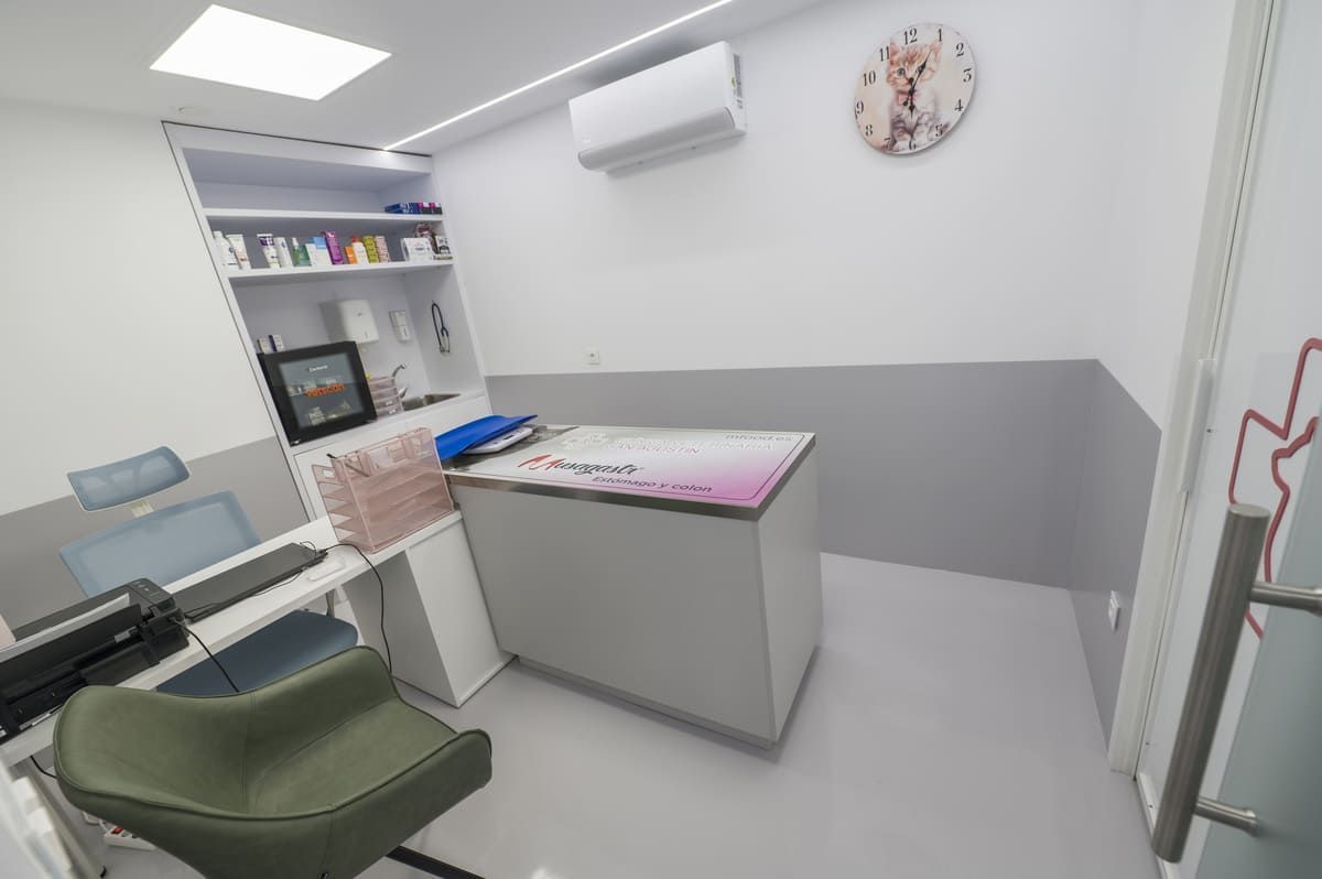 Sala de examen médico vacía con silla verde, mostrador, estantes y reloj de pared.