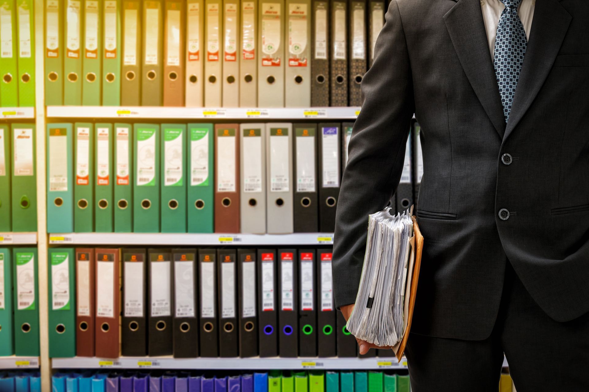 Un homme d'affaires tient des classeurs devant des étagères remplies de dossiers de bureau colorés.