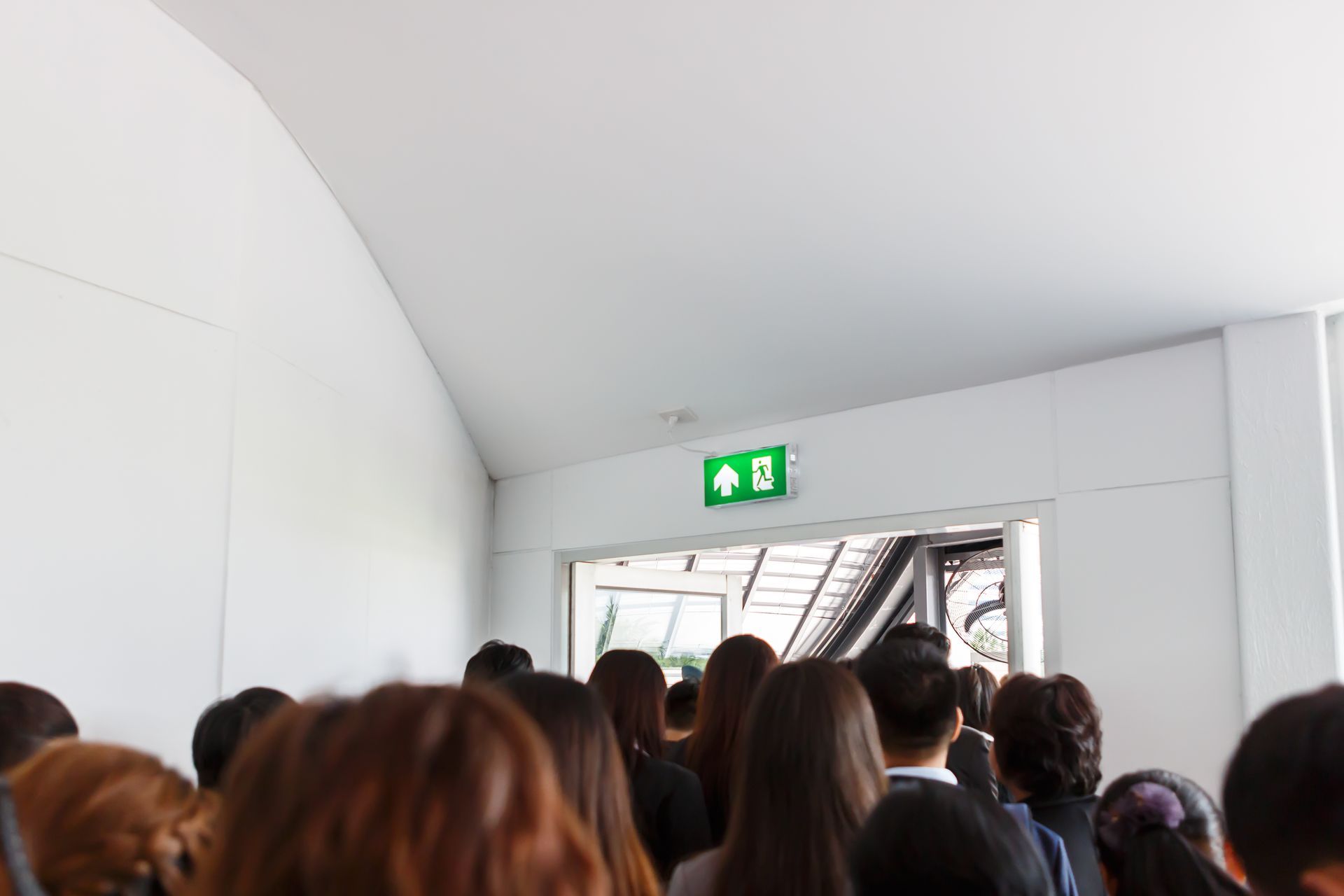 Couloir bondé, des gens faisant face à une porte ouverte sous un panneau de sortie vert.