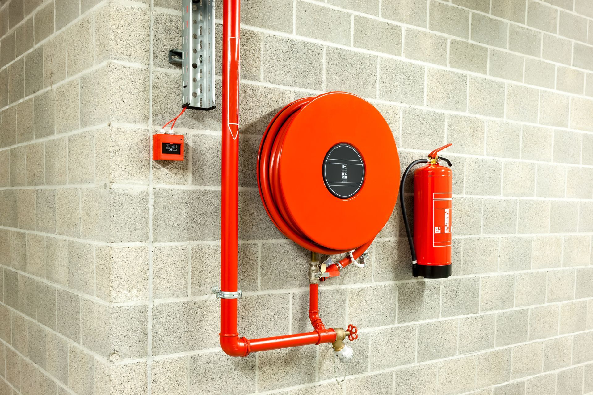 Dévidoir de tuyau d'incendie rouge et extincteur fixés sur un mur en parpaings gris.