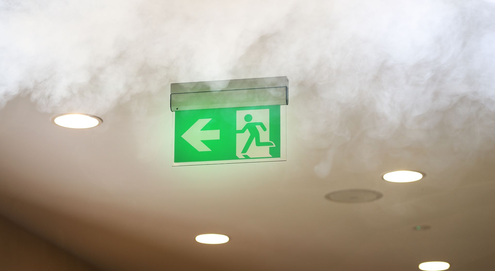 Panneau de sortie de secours vert avec flèche vers la gauche et silhouette courant, fixé au plafond au-dessus des lumières et de la fumée.