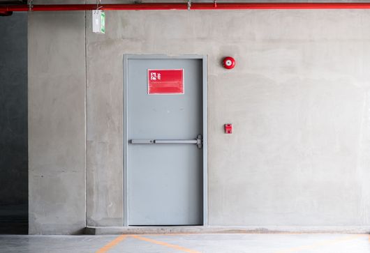 Porte métallique grise encastrée dans un mur en béton, avec des panneaux de sécurité rouges et un système d'alarme à proximité.