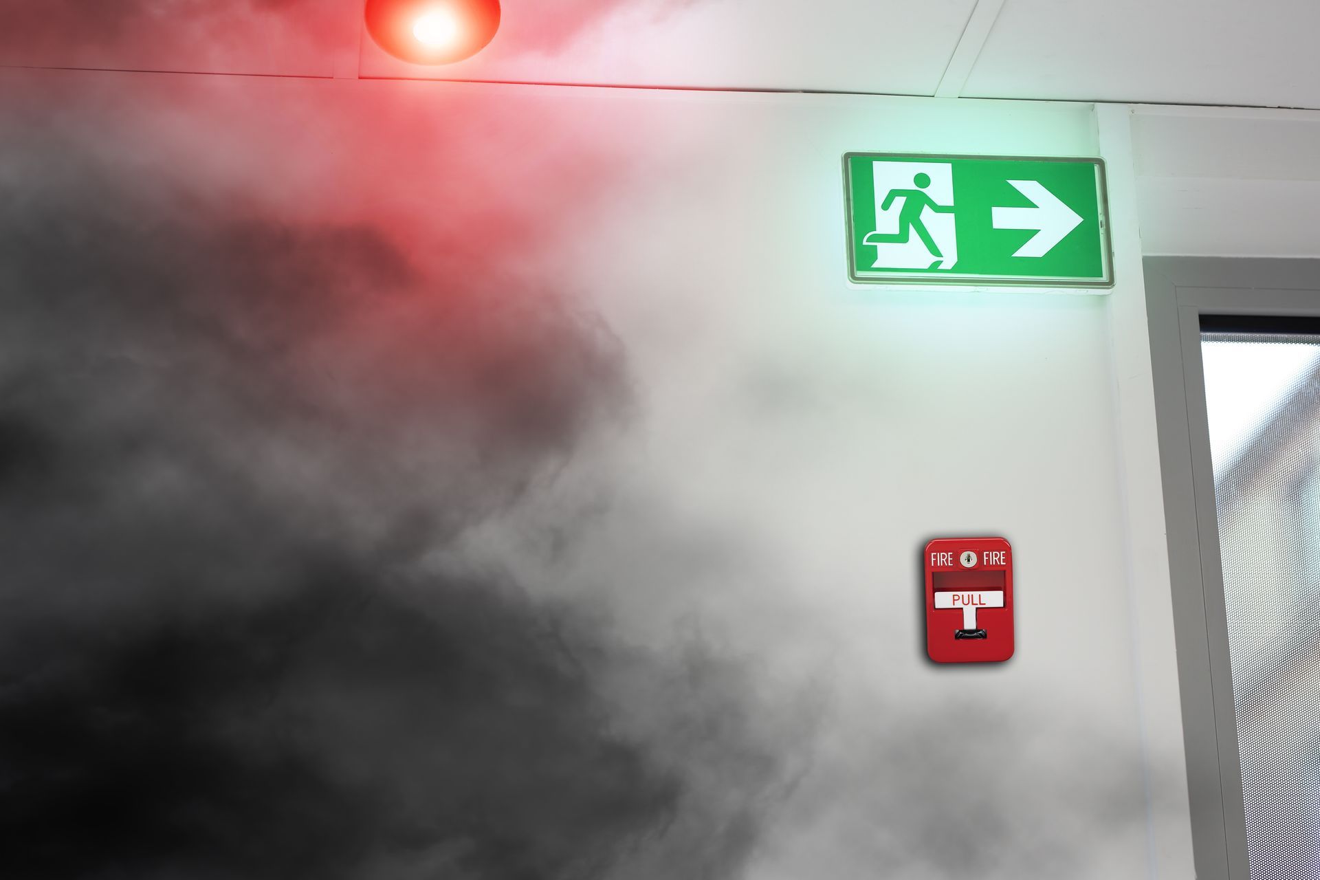 Couloir enfumé avec panneau de sortie vert lumineux et panneau d'alarme incendie rouge.