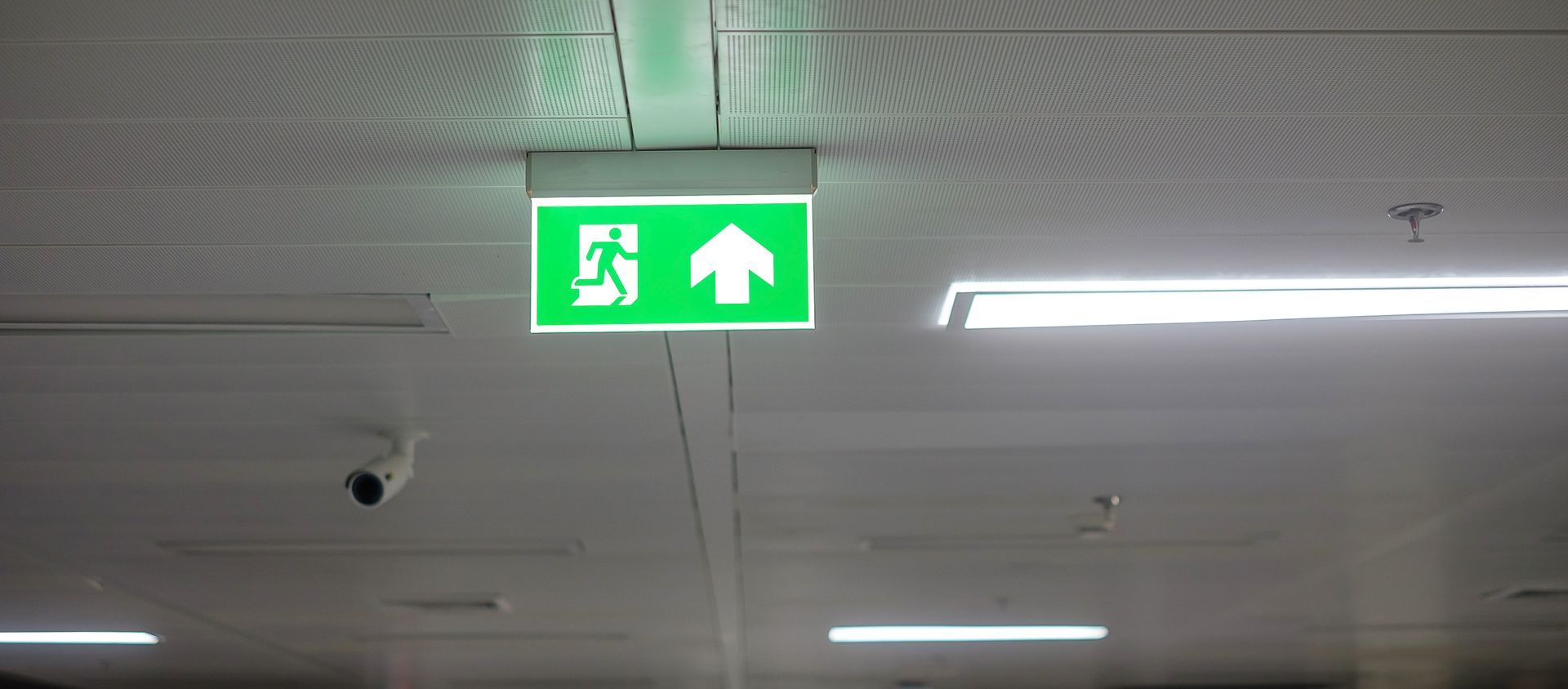 Panneau de sortie de secours vert avec une silhouette courant et une flèche pointant vers le haut, fixé au plafond à l'intérieur.