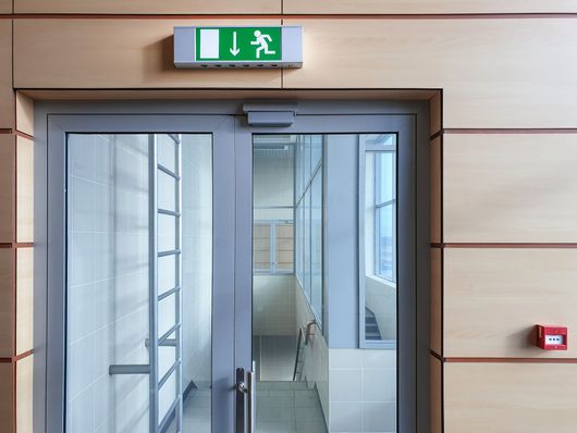 Porte de sortie vitrée avec panneau de sortie de secours vert au-dessus, dans un couloir moderne.