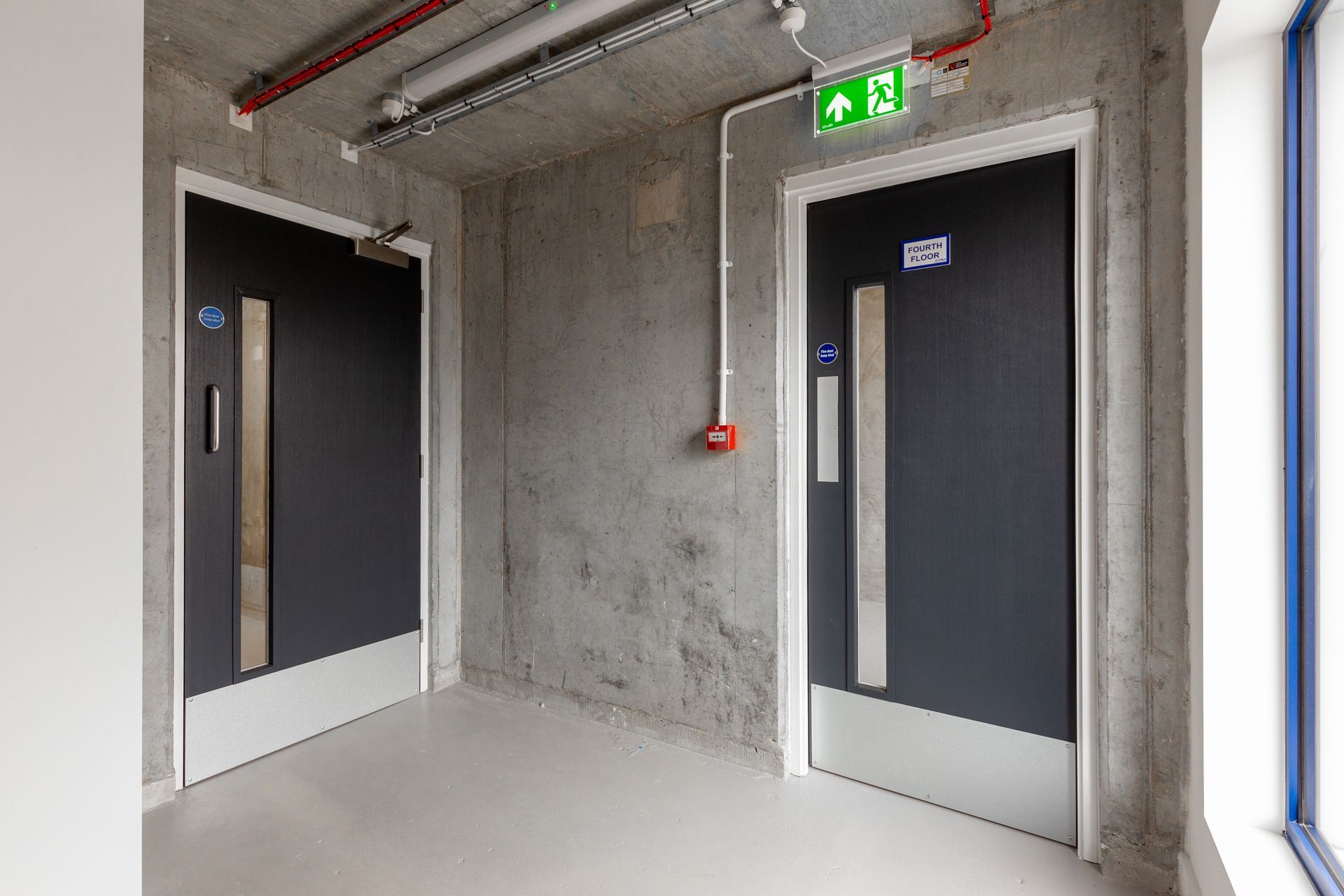 Couloir avec deux portes sombres, des murs en béton et un panneau de sortie vert au-dessus de la porte de droite.