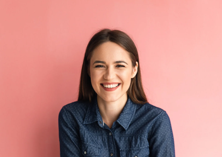 Une femme aux longs cheveux bruns sourit largement, vêtue d'une chemise en jean, sur un fond rose.